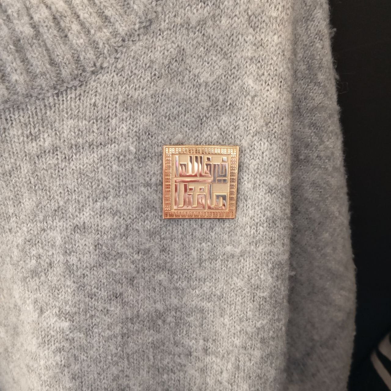 Rose gold square pin with Arabic text on a gray fabric background
