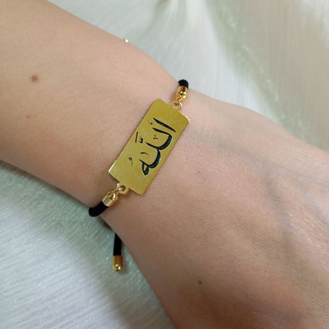 Gold bracelet with a rectangular tag featuring Arabic calligraphy on a wrist against a white background.
