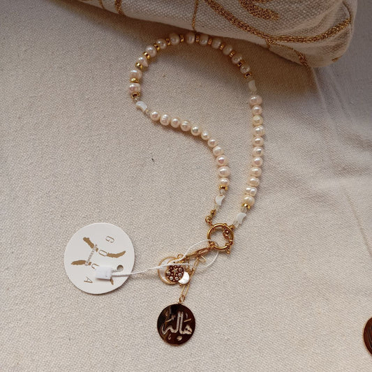 Pearl bracelet with gold accents on a textured beige surface, featuring a brand tag.