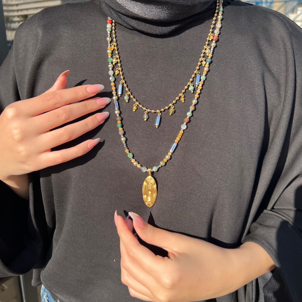 Person wearing a layered necklace with a gold pendant on a dark gray shirt.