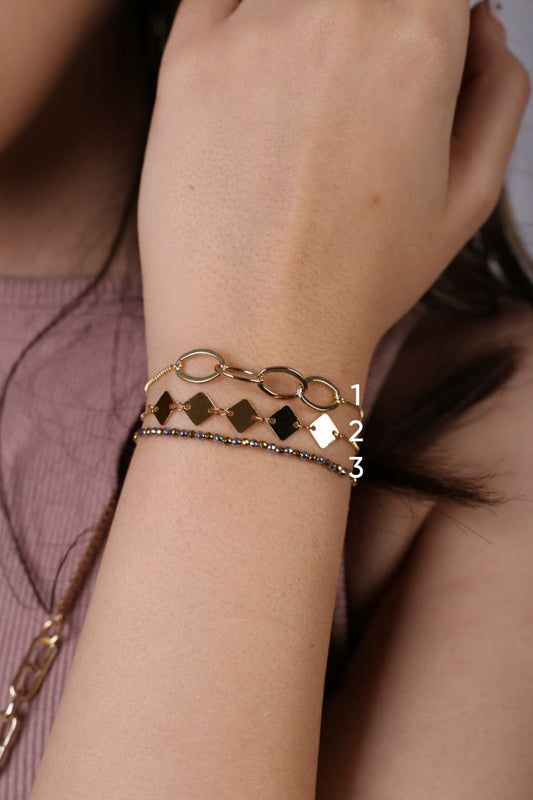 Close-up of a wrist wearing a gold bracelet with diamond-shaped charms on a blurred background.